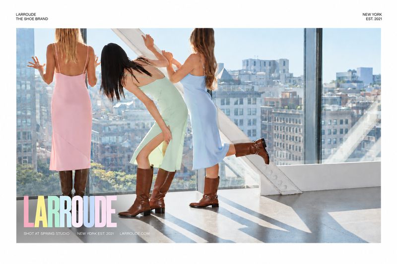 Three female models in pastel slip dresses and brown boots posing against large windows overlooking a city skyline.