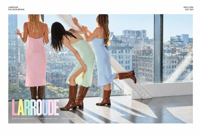 Three female models in pastel slip dresses and brown boots posing against large windows overlooking a city skyline.