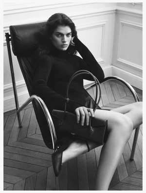 Monochrome fashion portrait of a model sitting in a modern leather armchair holding a dark handbag for Alaia.