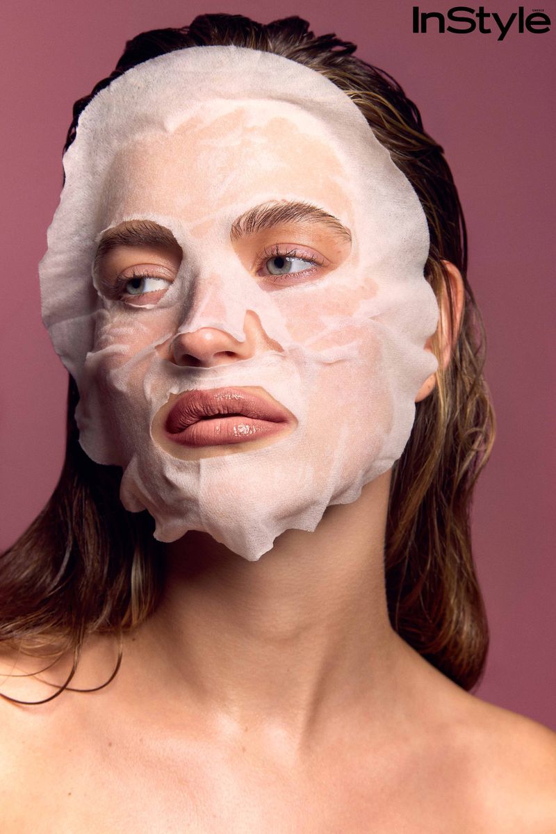 Close-up portrait of a woman wearing a white sheet face mask against a solid mauve background for InStyle Greece.