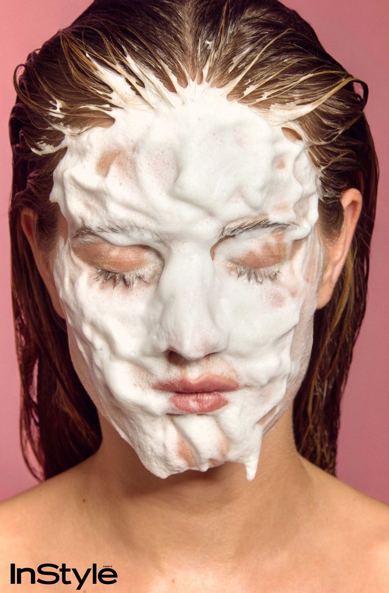 Close-up of a model with thick white cleansing foam covering her face and hair against a pink background for InStyle Greece skincare editorial.