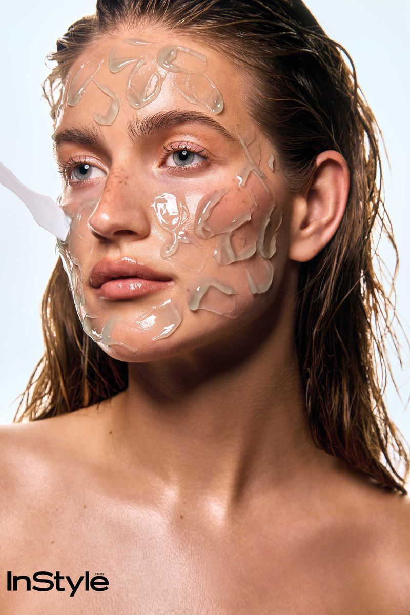 Close-up portrait of a woman with wet hair having skincare product applied to her face with a spatula against a white background.