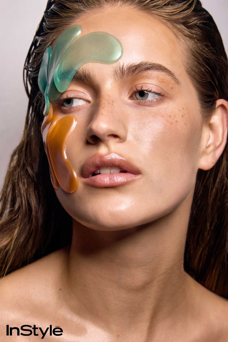 Close-up beauty portrait of a woman with wet hair and glossy skin wearing translucent green and amber skincare patches on her face.