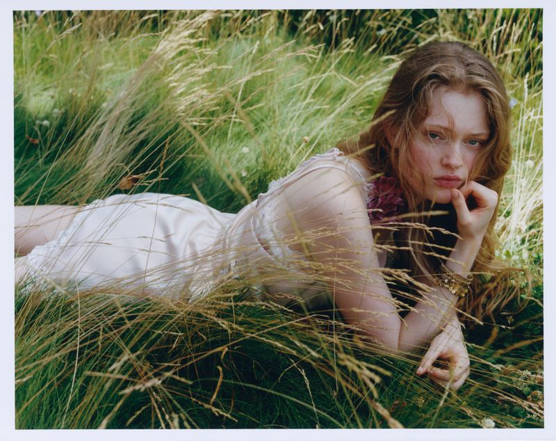Model lying in tall grass wearing a light-coloured dress and gold bracelets