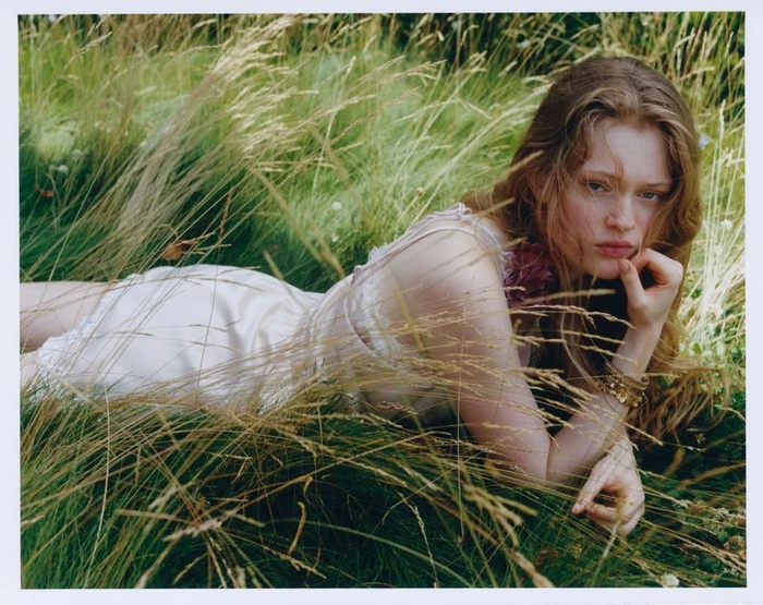 Model lying in tall grass wearing a light-coloured dress and gold bracelets