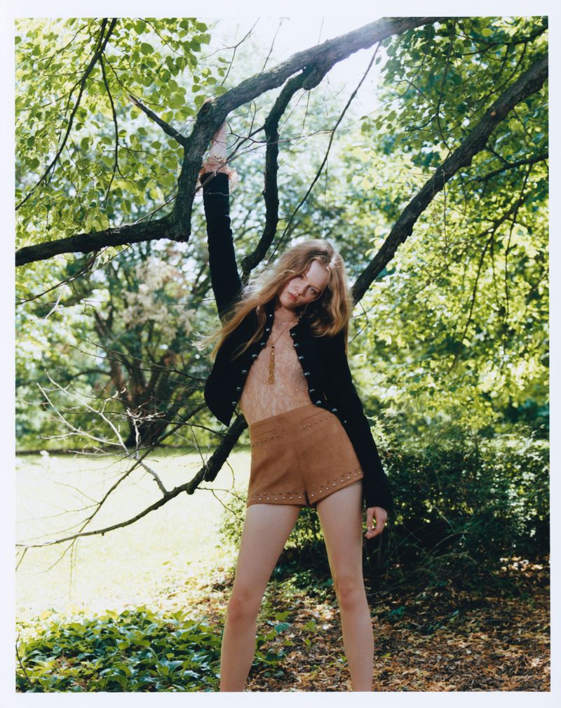 Model Ava Shipp holding onto a tree branch outdoors wearing a black jacket and brown shorts for Revolve editorial