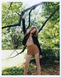 Model Ava Shipp holding onto a tree branch outdoors wearing a black jacket and brown shorts for Revolve editorial