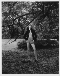 Model Ava Shipp in a dark jacket and shorts standing under leafy trees for a Revolve editorial photographed by Agata Serge.