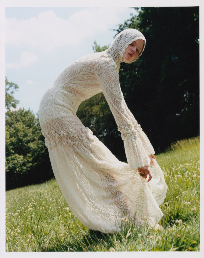 Model in a cream lace hooded dress bending forward in a grassy meadow with trees against an overcast sky.