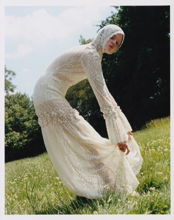 Model in a cream lace hooded dress bending forward in a grassy meadow with trees against an overcast sky.