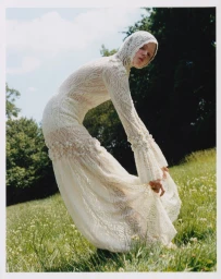 Model in a cream lace hooded dress bending forward in a grassy meadow with trees against an overcast sky.