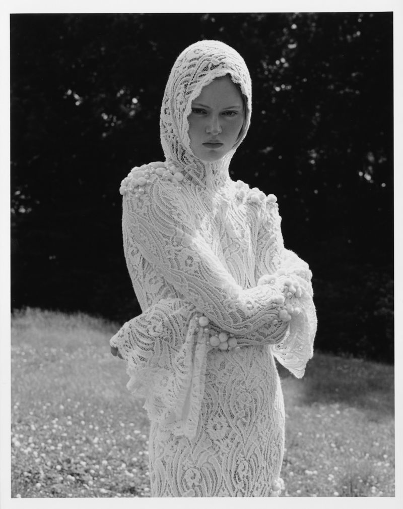 Model Ava Shipp in a white lace hooded dress posing outdoors in a grassy field for the Revolve Second Nature editorial.