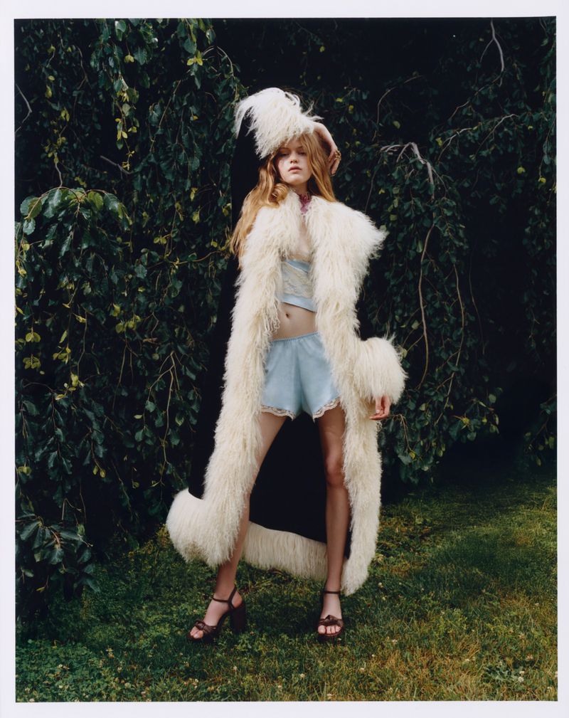 Model wearing a long white fluffy faux-fur coat and blue sleep set standing outdoors against dense foliage.