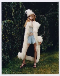 Model wearing a long white fluffy faux-fur coat and blue sleep set standing outdoors against dense foliage.