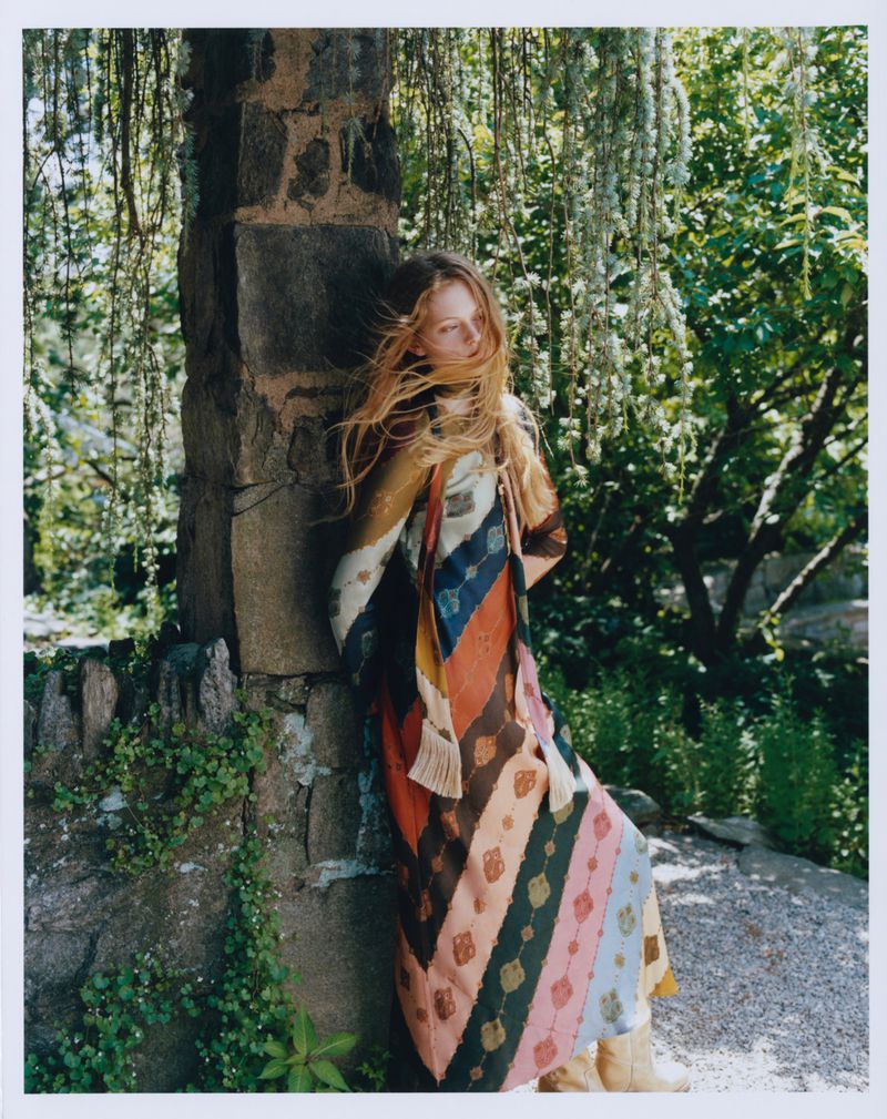 Model leaning against a stone pillar in a garden, wearing a striped patterned dress with cascading foliage above.