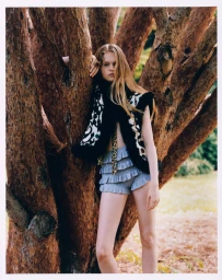 Model leaning against a large textured tree trunk wearing a patterned vest and ruffled shorts