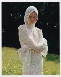 Model in a cream lace hooded dress standing outdoors against a green meadow and trees