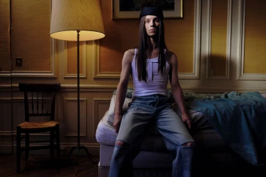 Model with long dark hair wearing a white tank top and ripped denim jeans sitting on a bed indoors