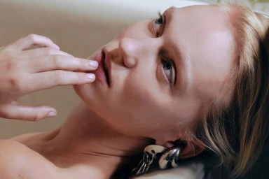 Close-up portrait of a woman with blonde hair wearing large silver chain earrings, touching her lips.