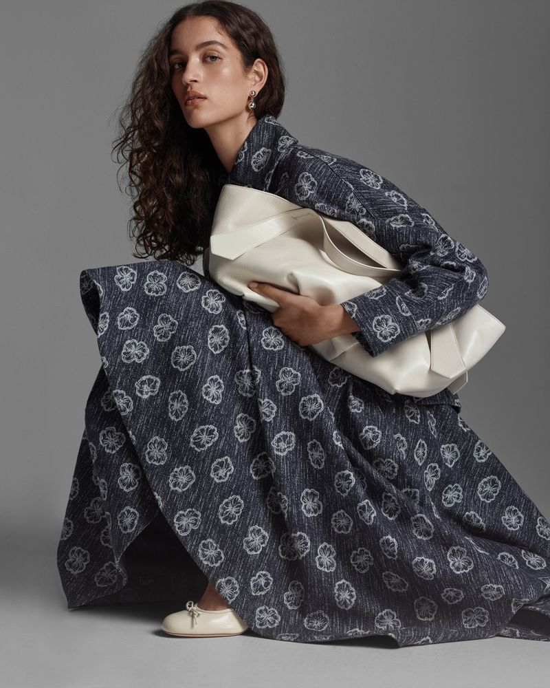 An adult woman is crouching indoors in a floral-patterned coat and dress, carrying a large white tote bag and wearing off-white shoes.