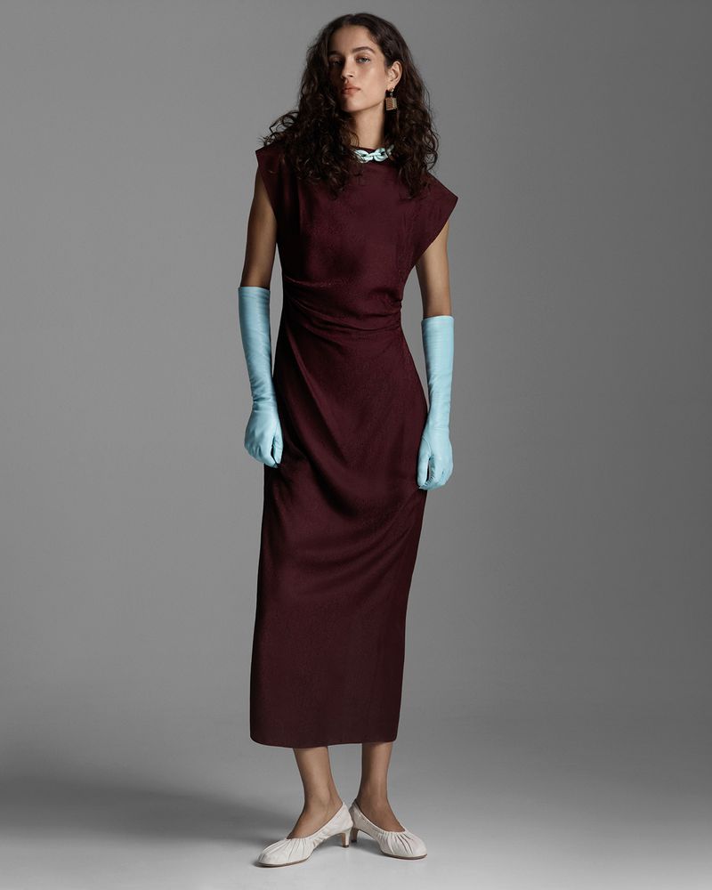 Adult female fashion model poses in a dark burgundy dress with pale blue gloves in a full shot against a plain gray studio background.