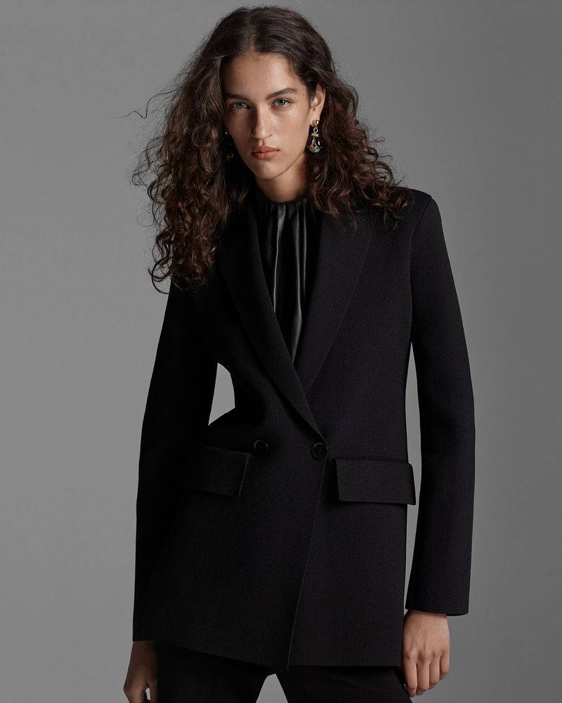 A woman with curly hair poses indoors wearing a black blazer and blouse. Fashionable dangling earrings accent the ensemble.