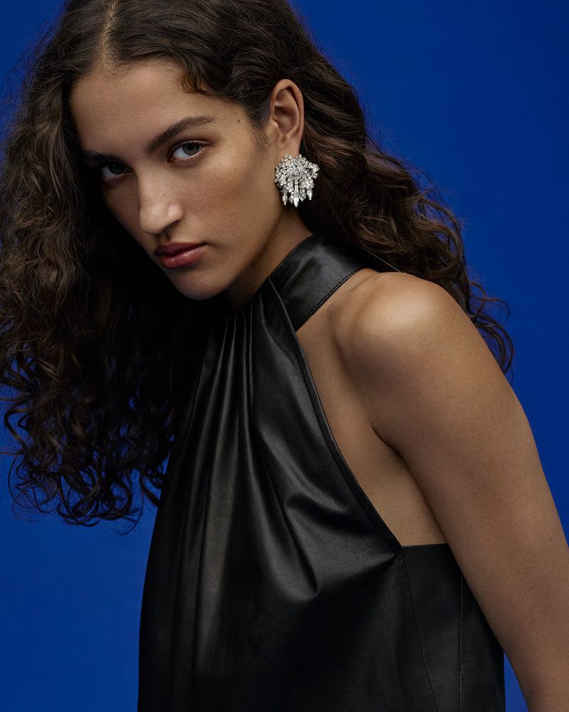 Portrait of a woman with dark wavy hair, wearing a black halter dress and large chandelier earrings, against a solid royal blue background.