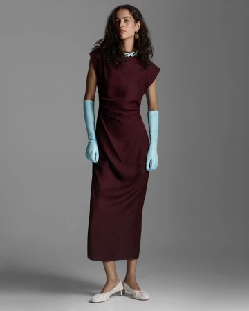 Model Lily Vander Meeden poses in a long burgundy column dress and pale blue opera gloves against a seamless grey backdrop.