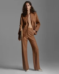 Model standing against a grey backdrop wearing a brown pinstripe suit jacket and matching trousers while holding a cream handbag.