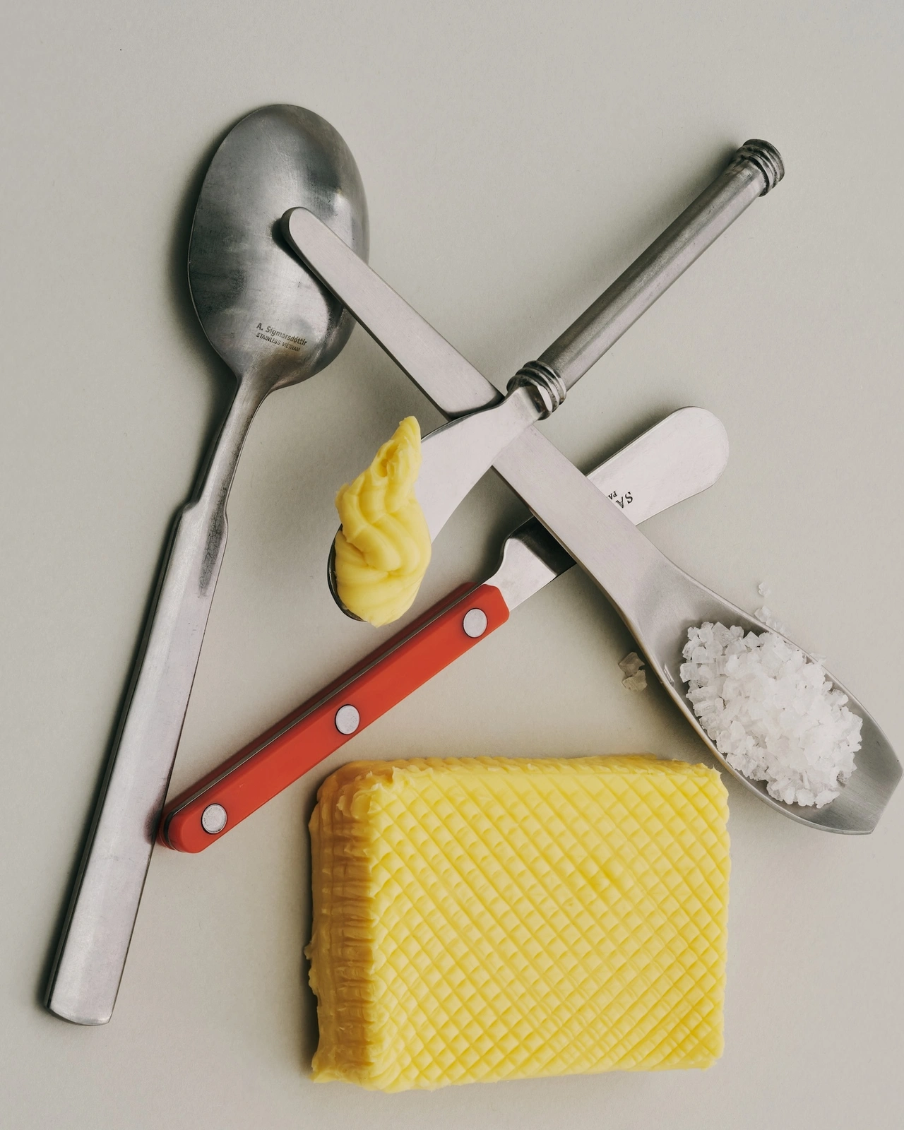 Flat lay of silverware, a textured block of butter, a dollop of yellow spread, and coarse salt against a pale background