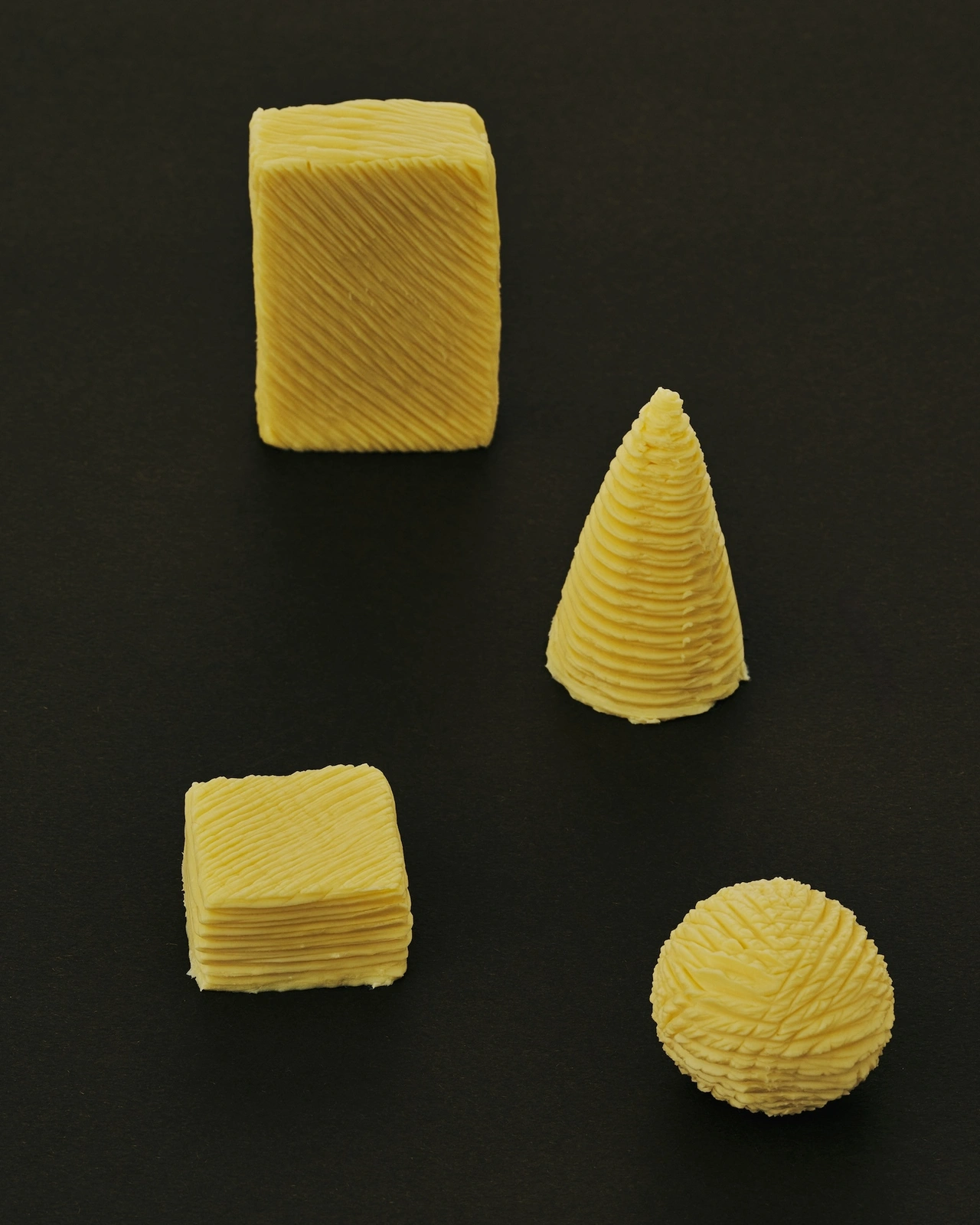 Four textured yellow geometric shapes including a cube, cylinder, square block, and sphere arranged on a dark background.
