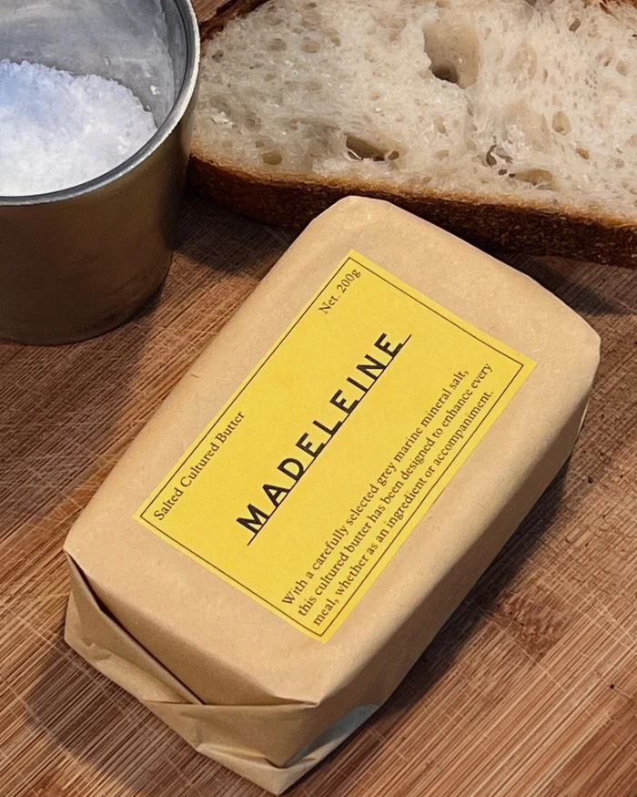 Packaged block of butter with a yellow label branded Madeleine, resting on a wooden surface beside bread and salt.