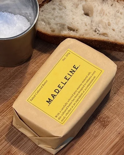 Packaged block of butter with a yellow label branded Madeleine, resting on a wooden surface beside bread and salt.