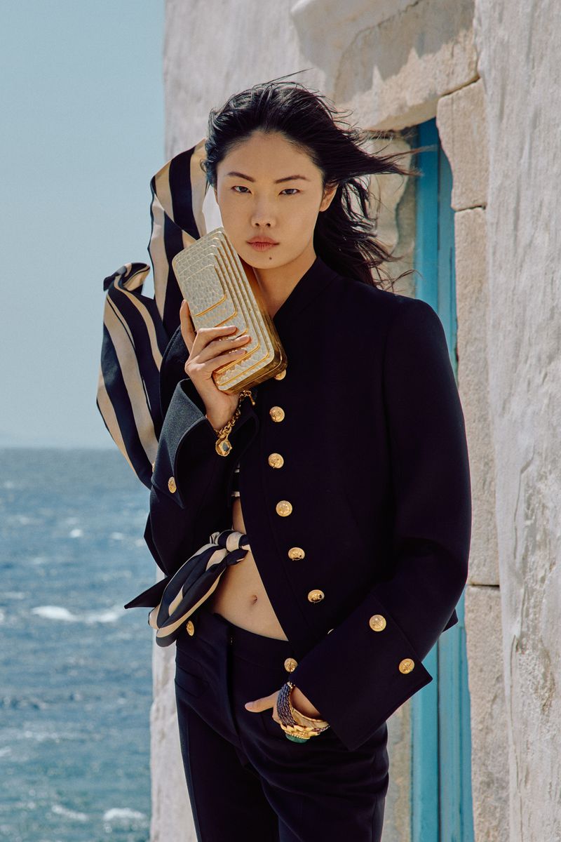 Woman in black suit poses holding gold clutch bag outdoors near building with ocean backdrop in a fashion editorial style shot.
