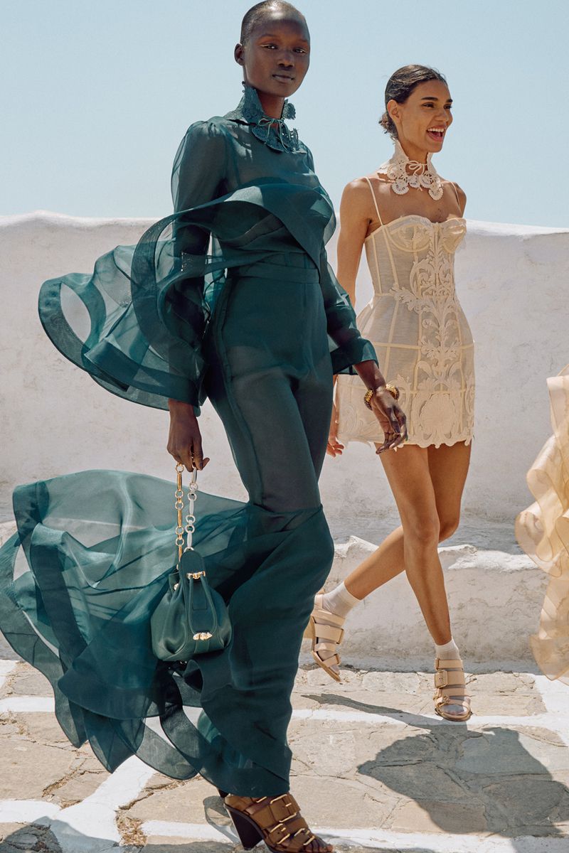 Two women in stylish dresses pose on white steps under a bright sky, showcasing flowing fabrics and elegant designs. 