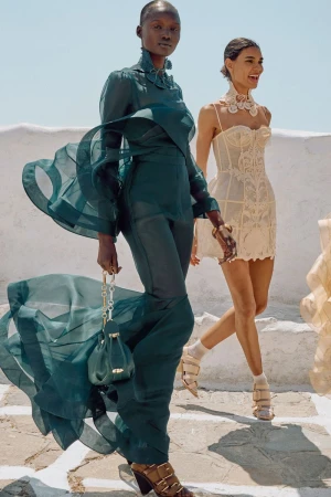 Two women in stylish dresses pose on white steps under a bright sky, showcasing flowing fabrics and elegant designs.