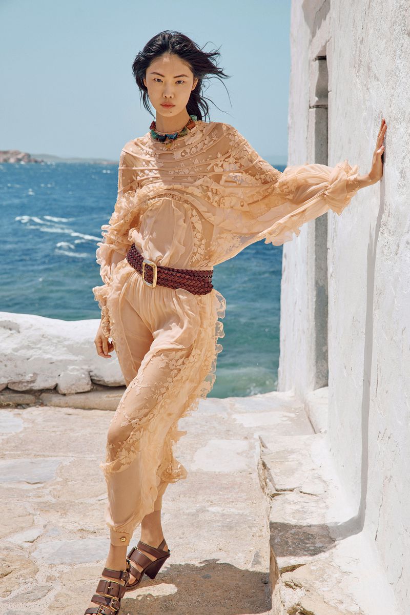 An Asian woman in a peach lace outfit and platform sandals poses outdoors by a white wall with the ocean visible behind her.