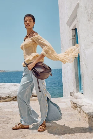 Model wearing a sheer off-the-shoulder ruffled top, wide-leg denim pants, and a brown leather bag on a sunny seaside terrace.