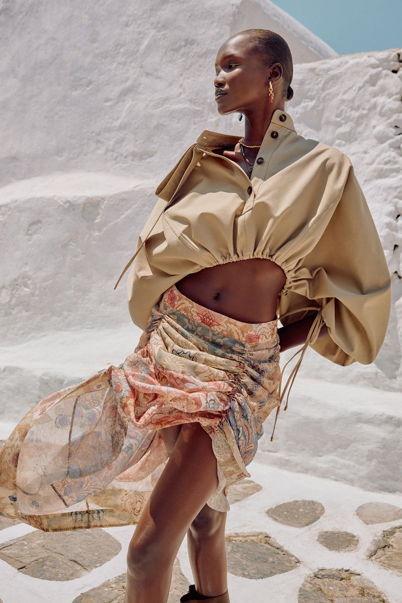 A slender Black woman poses outdoors in a tan top and patterned mini-skirt against a white stone wall, showcasing fashion and style.