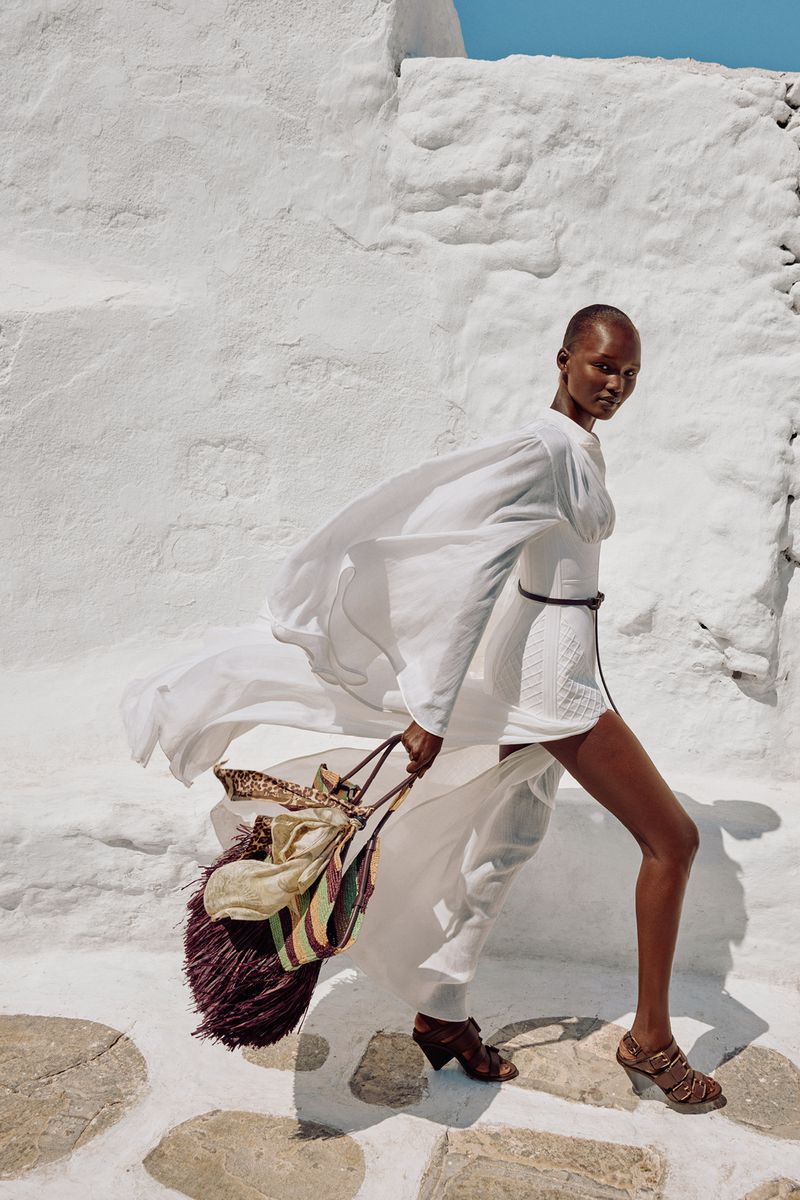 Woman in white dress and brown heels carrying fringed bag walking on stone path with whitewashed walls in outdoor fashion shot.