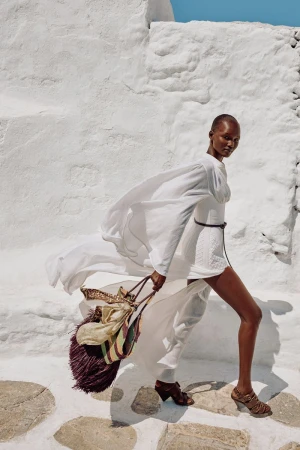 Woman in white dress and brown heels carrying fringed bag walking on stone path with whitewashed walls in outdoor fashion shot.
