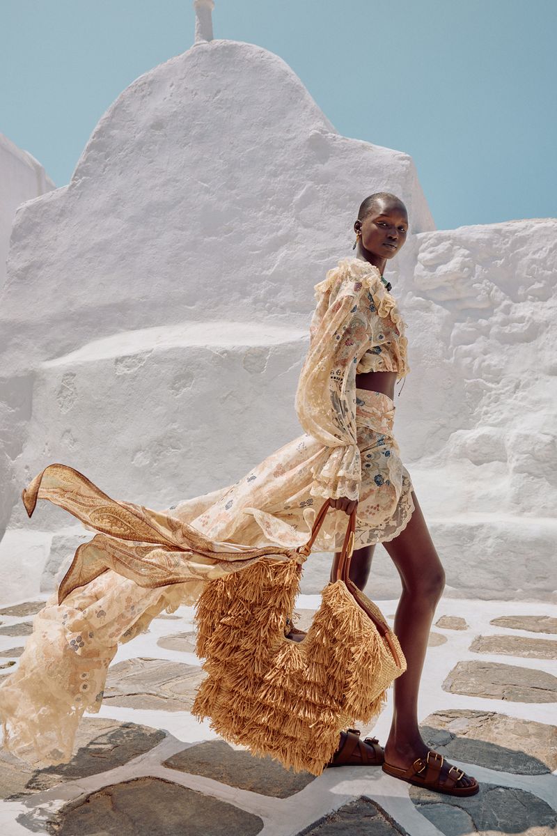An adult woman wearing a floral two-piece outfit walks along a cobblestone path carrying a large straw fringe purse.
