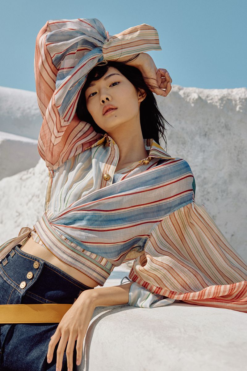 Portrait of a young woman in a striped shirt and denim jeans posing in front of a white wall and blue sky.