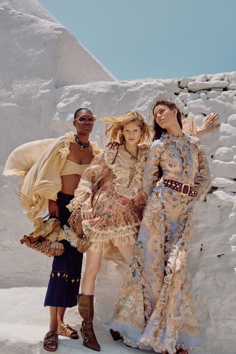 Three women pose outdoors in flowing dresses against a white, textured wall. The dresses feature pastel colors, lace, and ruffles. 