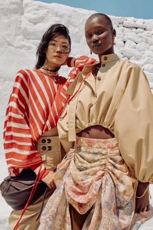 Two models pose in outdoor fashion shot with bold stripes and a draped skirt, against a white background, showcasing modern, high-end apparel.