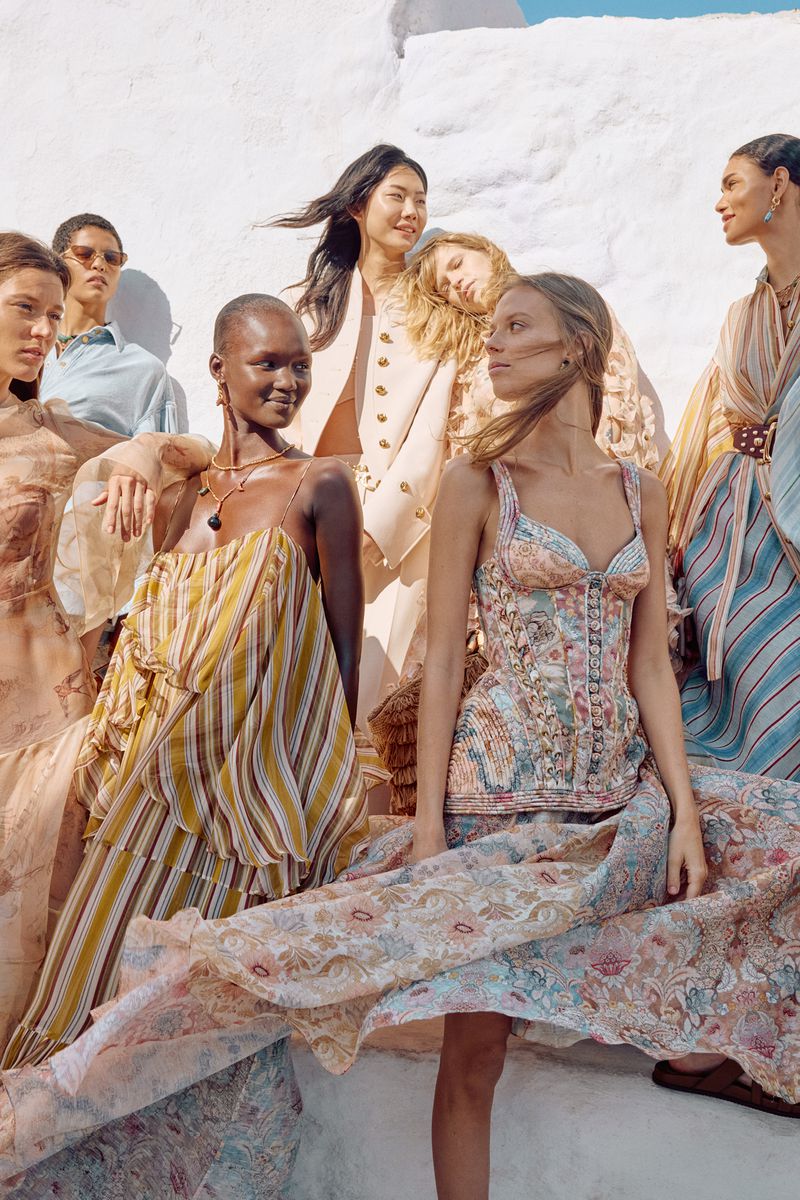 Group of diverse women in flowing dresses and neutral tones, posing against a bright white wall in a fashion editorial style shoot.