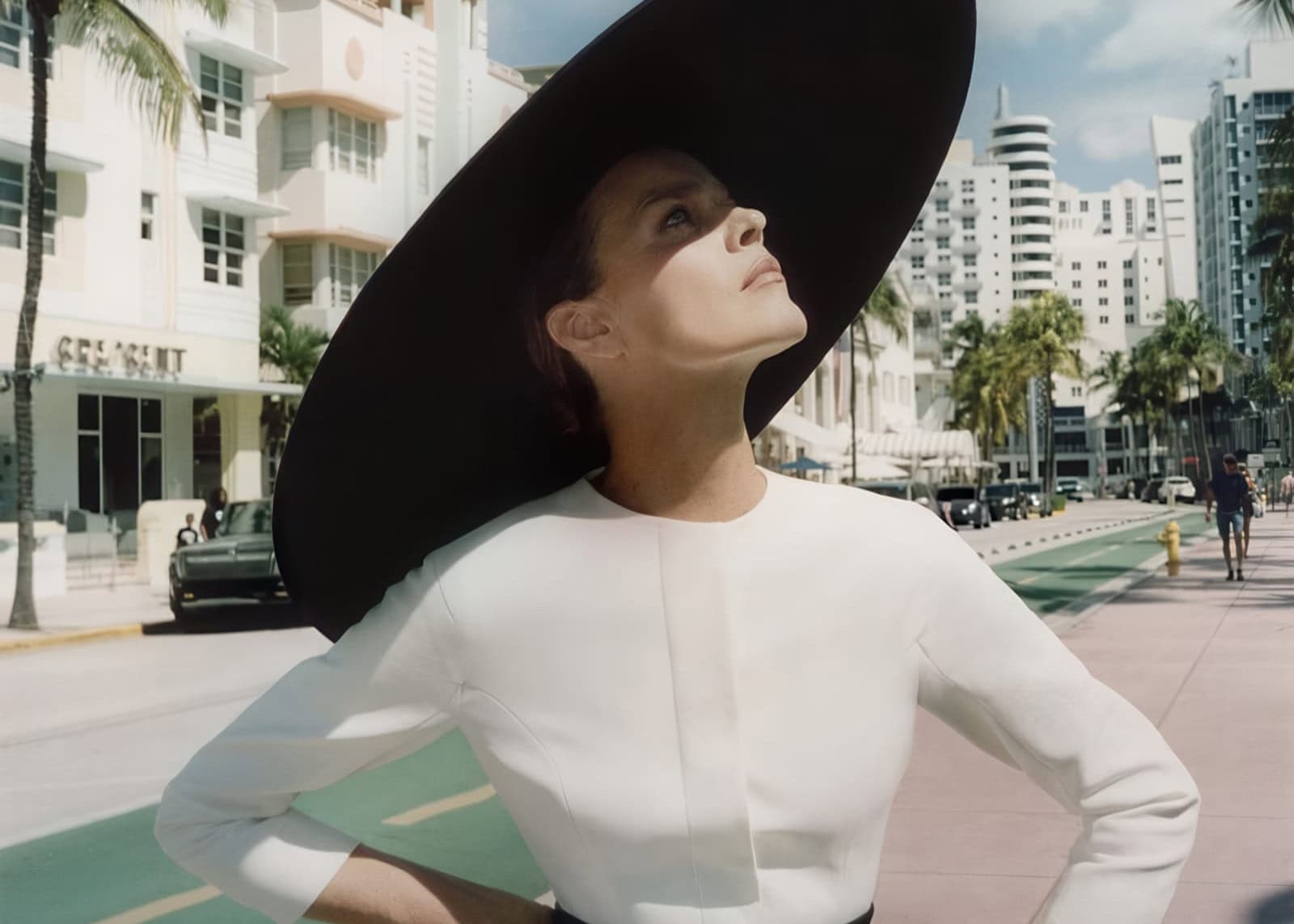 Eva Herzigova in a wide black sun hat and white top outdoors on a sunny street