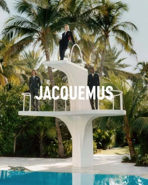 Three models in dark suits on a white concrete diving platform beside a pool, surrounded by lush palm trees.