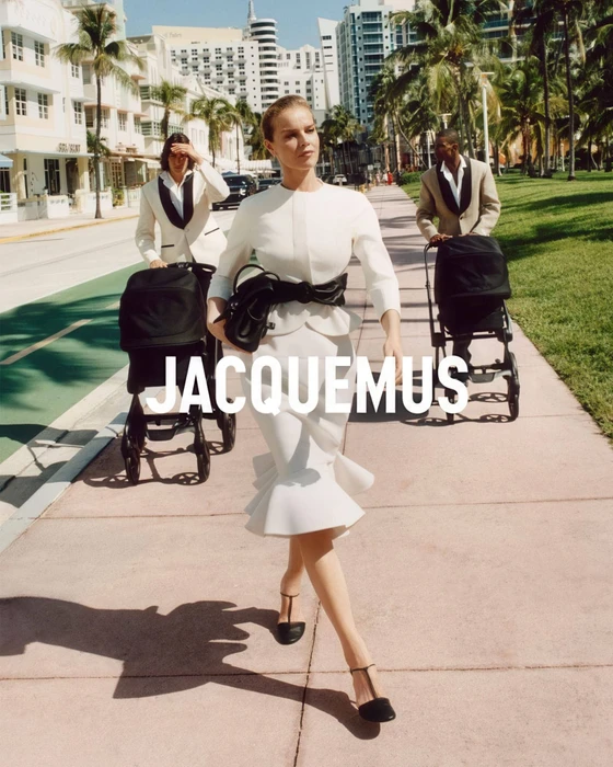 Eva Herzigova walking along a palm-lined sidewalk pushing a pram, wearing a structured white dress and black belt for Jacquemus.