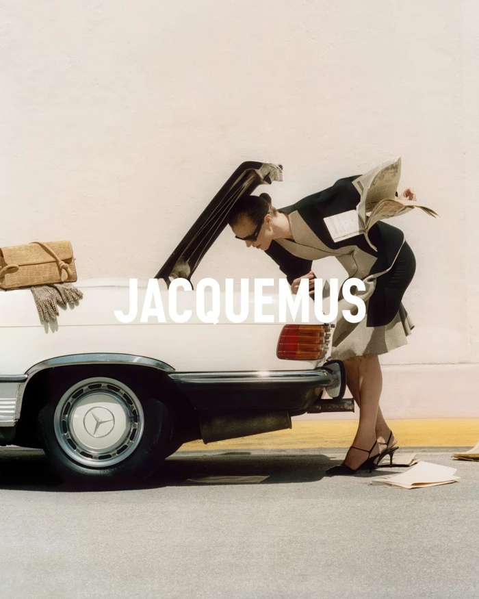 Model leaning into the open trunk of a white vintage car while holding newspapers, styled for the Jacquemus Le Turismo campaign.
